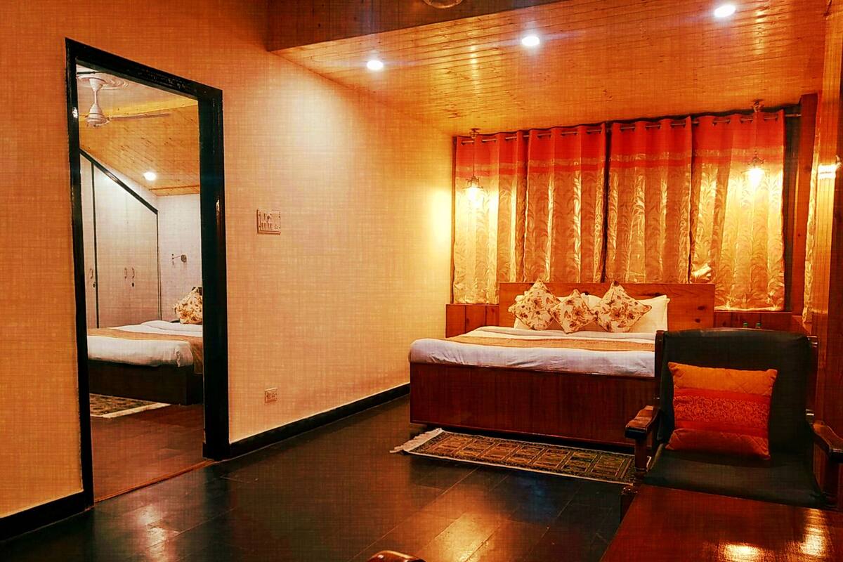 A hotel room with a wooden floor, a bed with floral pillows, a chair, and a mirror reflecting the room.