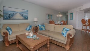Un salón con un sofá color beige, una mesa de centro de madera y un cuadro de un muelle.