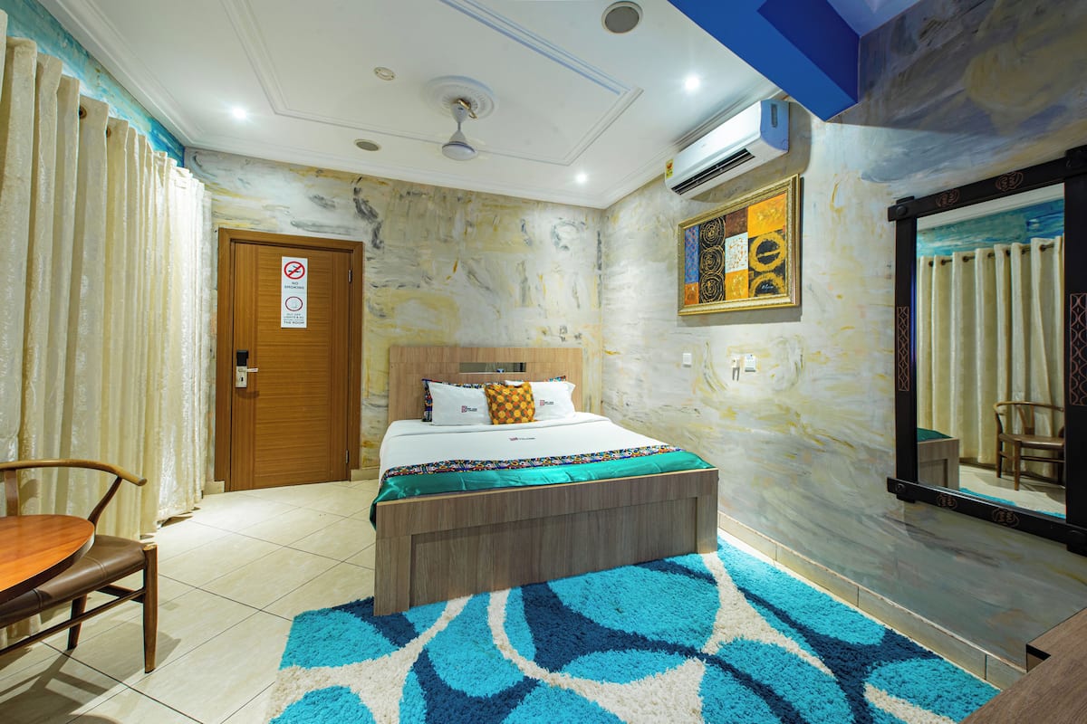 Une chambre d’hôtel avec un lit, une chaise en bois, un tapis à motifs bleus et blancs, une porte en bois et un tableau au mur.