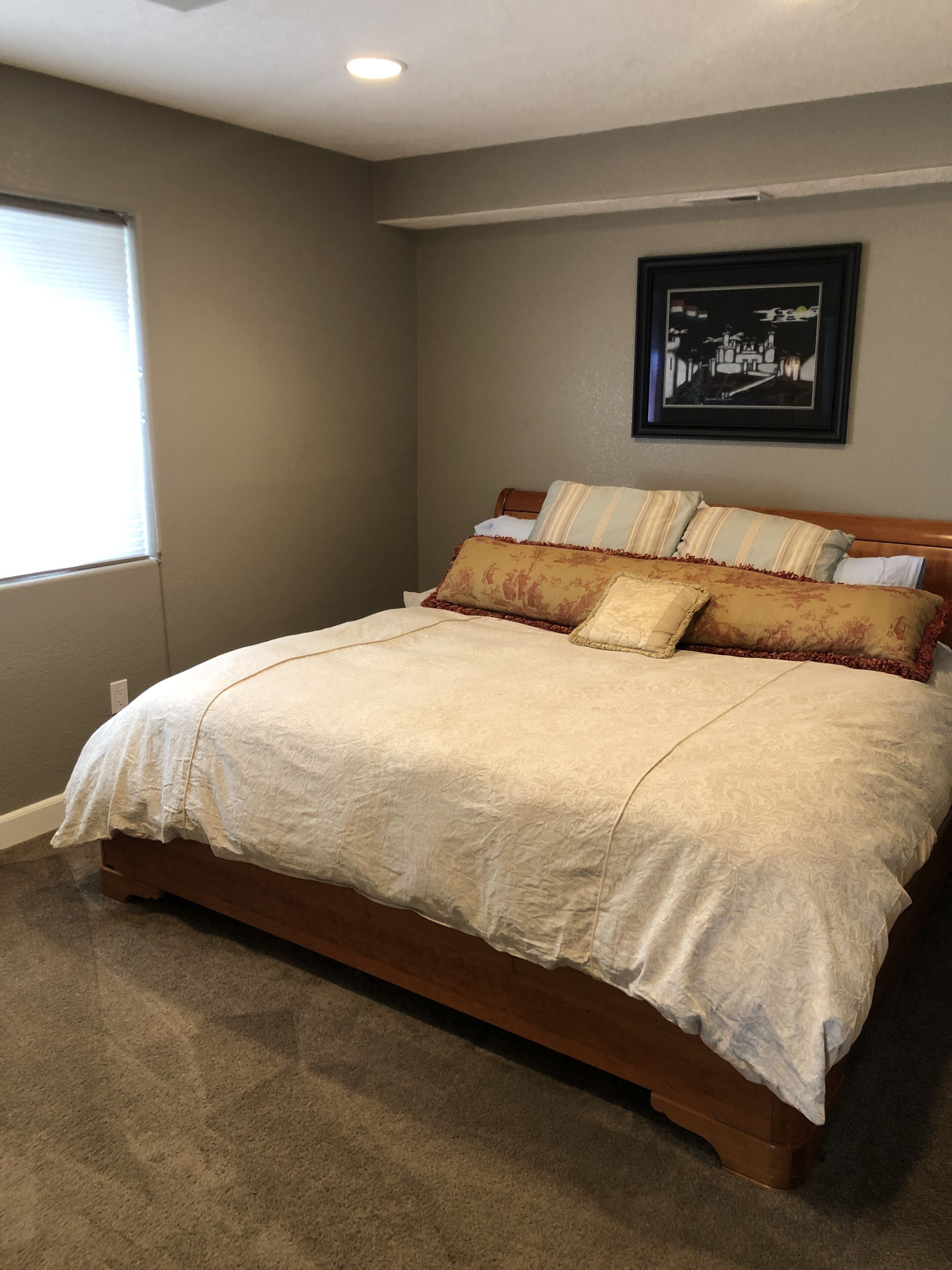 A bedroom with a large bed, a window with blinds, a framed picture on the wall, and a carpeted floor.