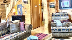 Ein Holzblockbau-Innenraum mit Schlafzimmer im Obergeschoss, einer Couch, einem Sessel und einem Esstisch.