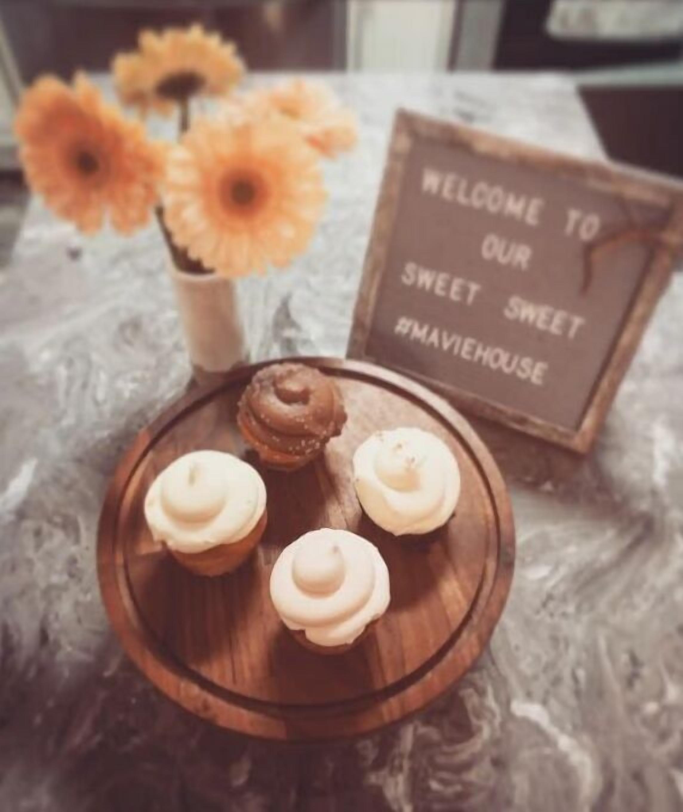 A wooden tray with four cupcakes on a marble surface, a sign that reads "WELCOME TO OUR SWEET #MAVEEHOUSE," and a vase with yellow flowers in the background.