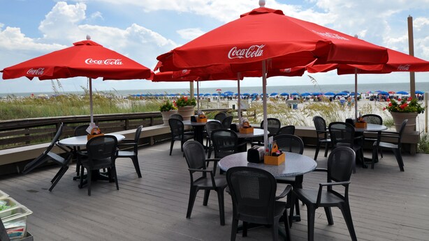 Uma área de estar externa com mesas e cadeiras sob guarda-sóis vermelhos da Coca-Cola, com vista para uma praia.