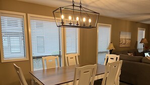 Uma área de refeições com mesa e cadeiras de madeira, um lustre e grandes janelas com persianas.