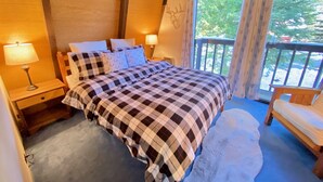 A cozy bedroom with a checkered bedspread, two bedside lamps, a wooden headboard, a window with curtains, and a balcony chair. - Cozy family friendly cabin  (Truckee)