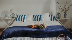 A neatly made bed with a blue and white striped pillowcase, a basket of fruits, and a vase with branches on a bedside table.
