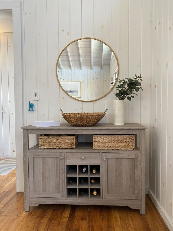 A grey wooden cabinet with a round mirror, two woven baskets, and a vase with green plants. - The Raabbit Hole (Ocean Beach)