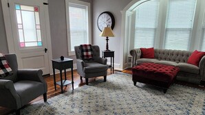 Una sala de estar con un sillón gris, un sofá con cojines rojos, un tapete estampado y un reloj en la pared.