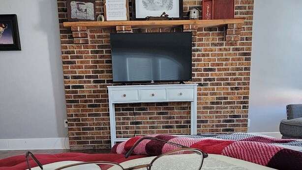 Una habitación con chimenea de ladrillo, un televisor sobre un mueble bajo y un libro abierto con gafas encima.