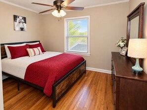 Une chambre à coucher avec un lit, un ventilateur de plafond, une fenêtre avec des stores, une commode en bois et une lampe.