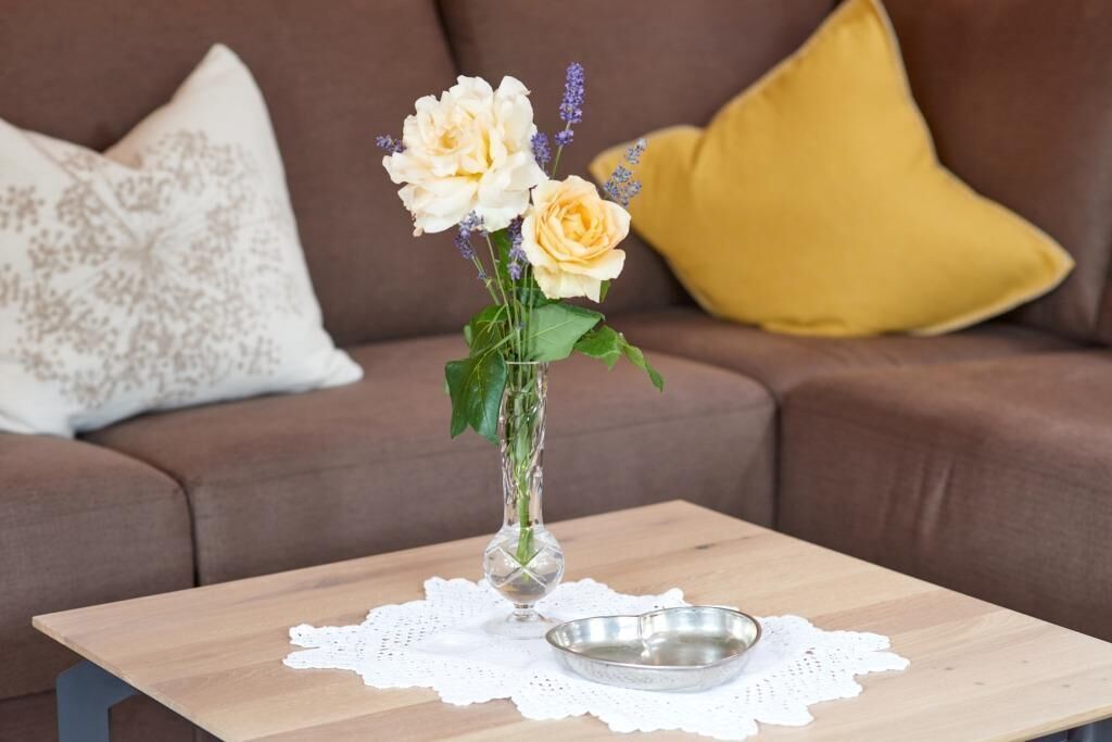 A clear view of a vase with flowers on a wooden table, a metal dish, and a lace doily.