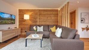 A modern living room with a wooden wall panel, a sofa, a coffee table, and a TV on the left. - (06) Comfort vacation apartment with south-east-facing balcony, approx. 67 sqm (Reit im Winkl)
