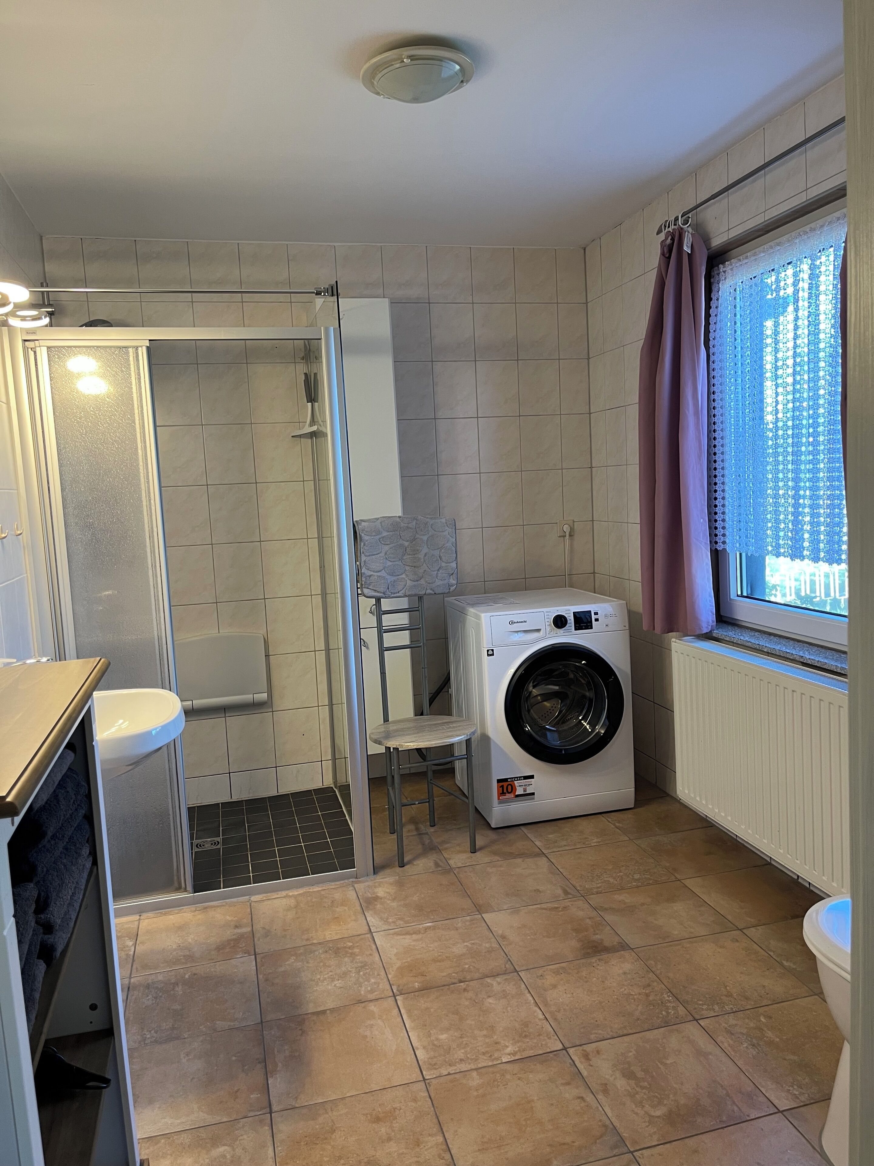 Une petite buanderie comprenant un lave-linge, un petit tabouret, un espace douche et une fenêtre avec des rideaux.