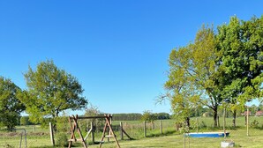 A grassy outdoor area with a wooden swing set, a small blue container, and a fenced field. - Eldehof, Oldag family (Wredenhagen)