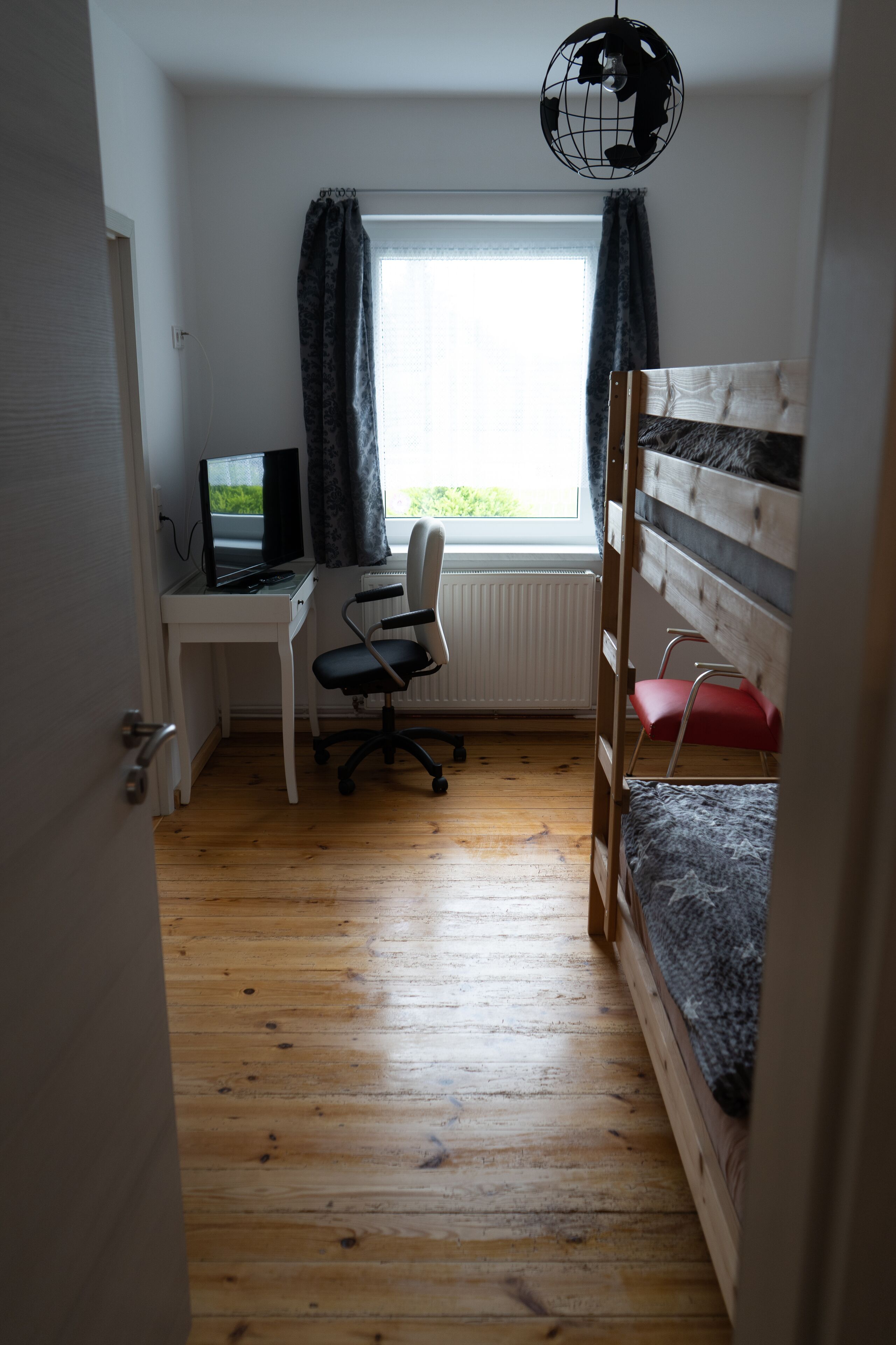 Ein Zimmer mit einem Etagenbett, einem Schreibtisch mit Computer, einem Stuhl und einem Fenster mit Vorhängen.