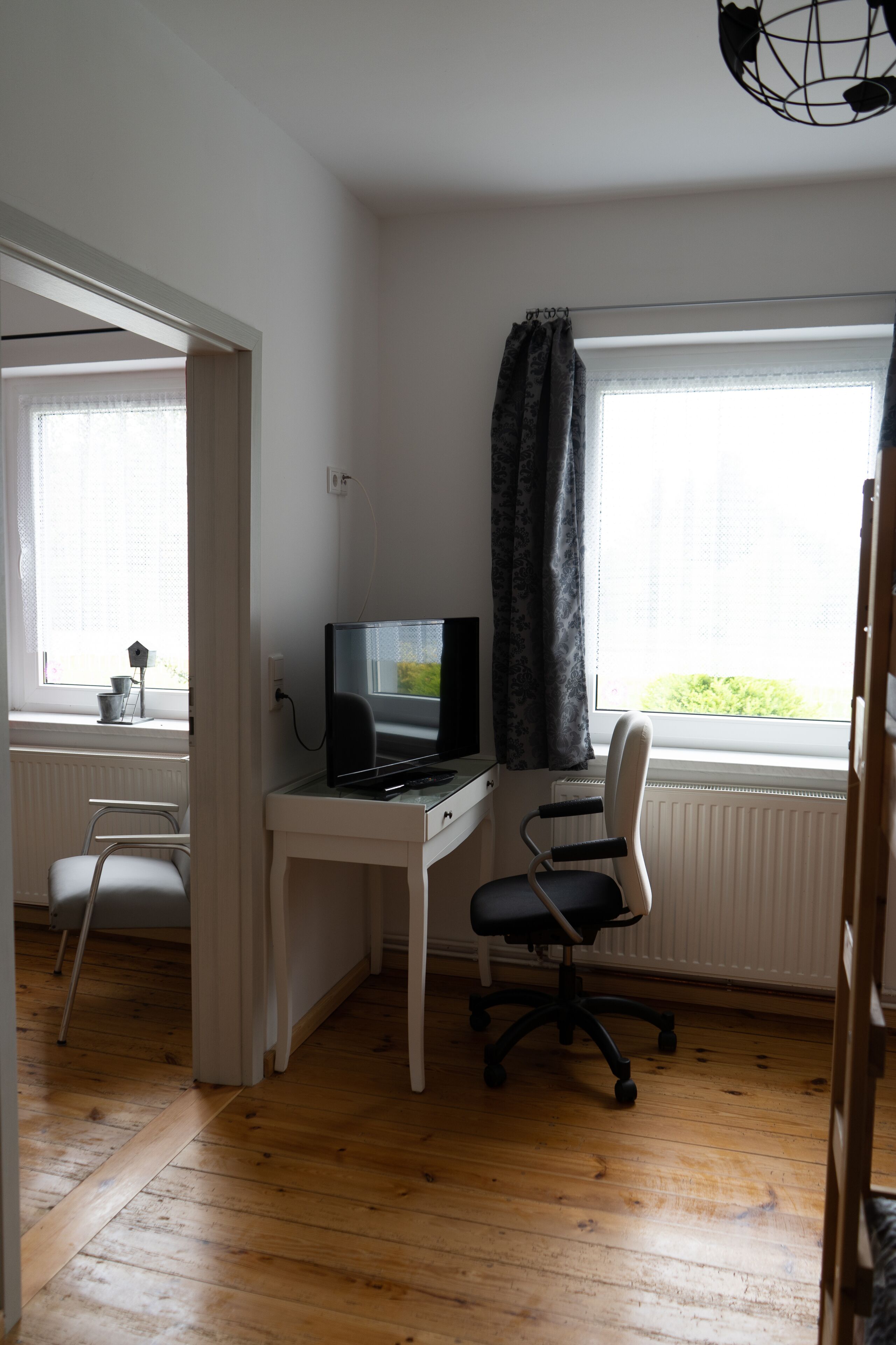Een kleine, goed verlichte kamer met een houten vloer, een bureau met een computer en een stoel, een raam met gordijnen en een radiator.