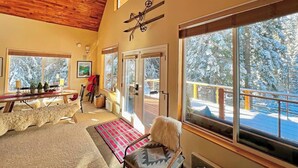A cozy room with a wooden ceiling, large windows, a ski-themed wall decoration, a bench, and a view of a snowy deck outside.