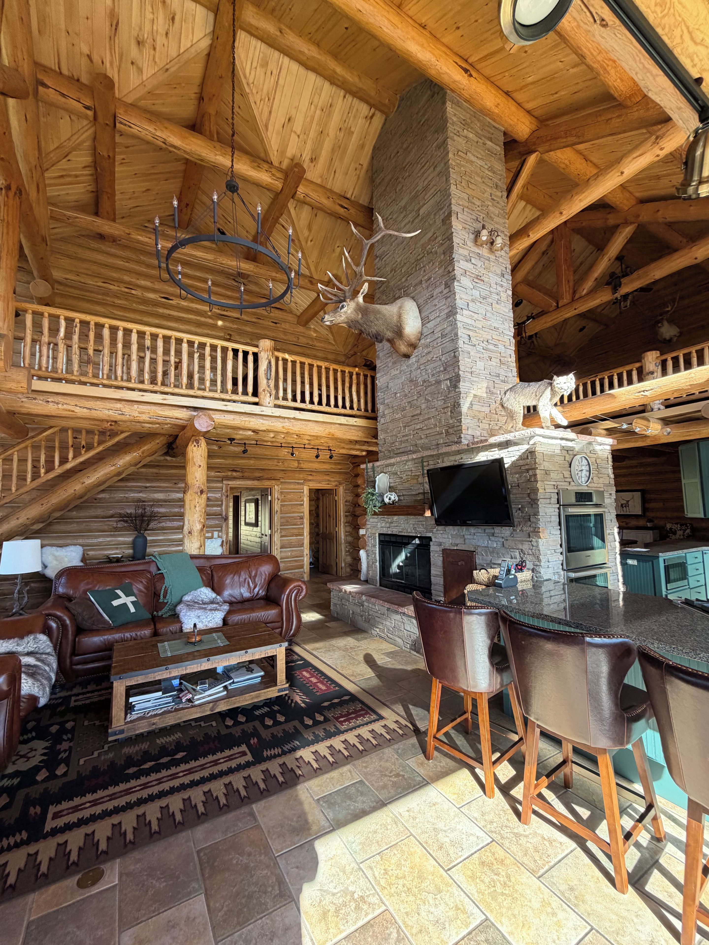 A log cabin interior with a stone fireplace, leather furniture, and a mounted deer head.