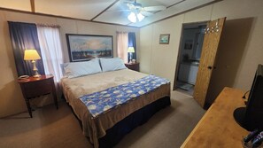 Habitación de hotel con cama, dos lámparas de noche, ventilador de techo, un cuadro en la pared y vista a la cocina a través de una puerta abierta.