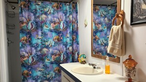 Un baño con cortina de ducha estampada con motivos de peces, un lavamanos con espejo, una toalla colgada en la pared y un pequeño recipiente con conchas marinas.