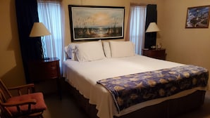 Una cama bien hecha con una colcha blanca y un edredón con estampado azul y blanco. A la izquierda de la cama hay una silla de madera, una mesita de noche con una lámpara y un cuadro en la pared sobre la cama.