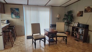Una sala de estar con piso de madera en patrón de espiga, una mesa de comedor con cuatro sillas, una estantería, una lámpara y un ventanal con persianas.