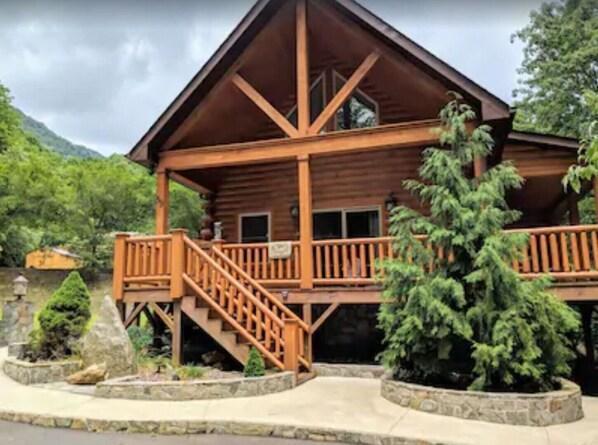 A wooden lodge with a balcony and stairs, surrounded by greenery and mountains.