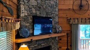A rustic interior with a stone fireplace, wooden walls, a mounted television, and a window with blinds. - "Rushing Bear!"   Fully furnished two bedroom cozy cabin you want to capture!" (Maggie Valley)