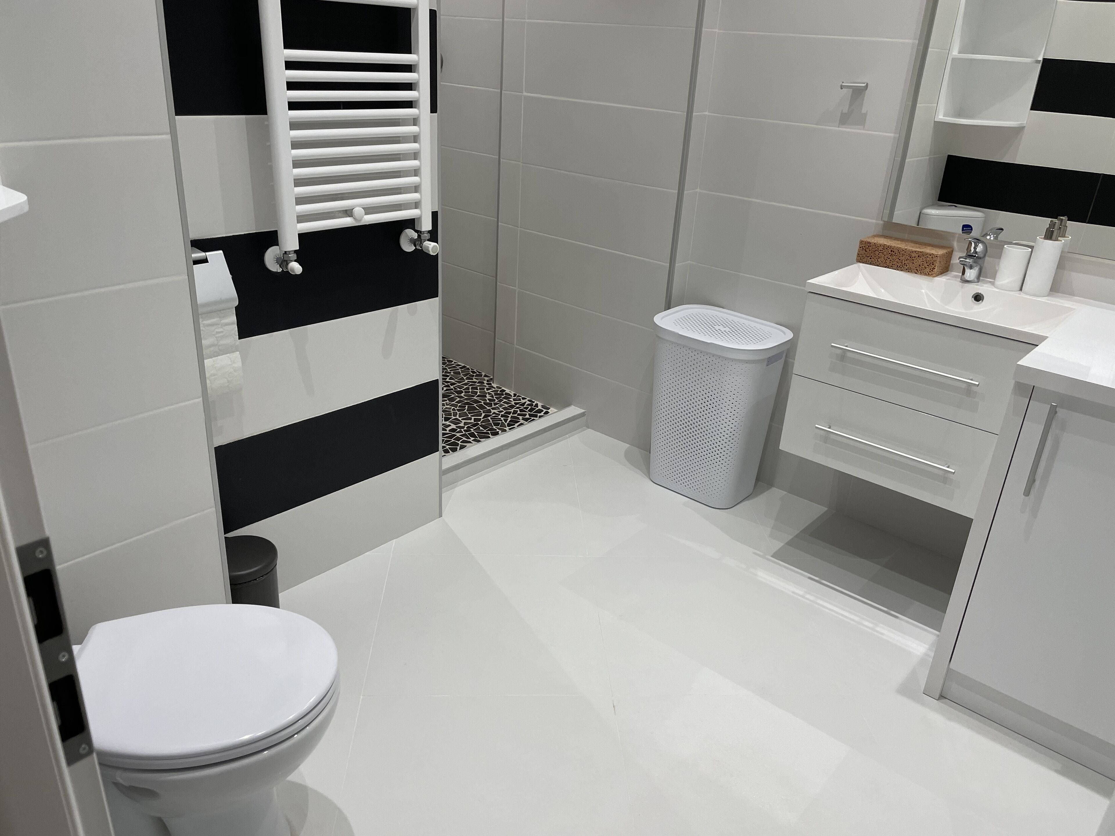 Een moderne badkamer met een toilet, een douche met een zwart-wit betegelde wand, een witte wastafel met spiegel en een prullenbak.