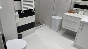 Een moderne badkamer met een toilet, een douche met een zwart-wit betegelde wand, een witte wastafel met spiegel en een prullenbak.
