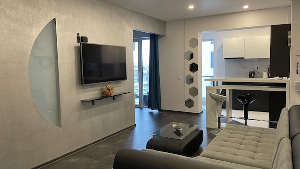 A modern living room with a flat-screen TV, a sofa, a coffee table, and a dining area with a kitchen in the background.