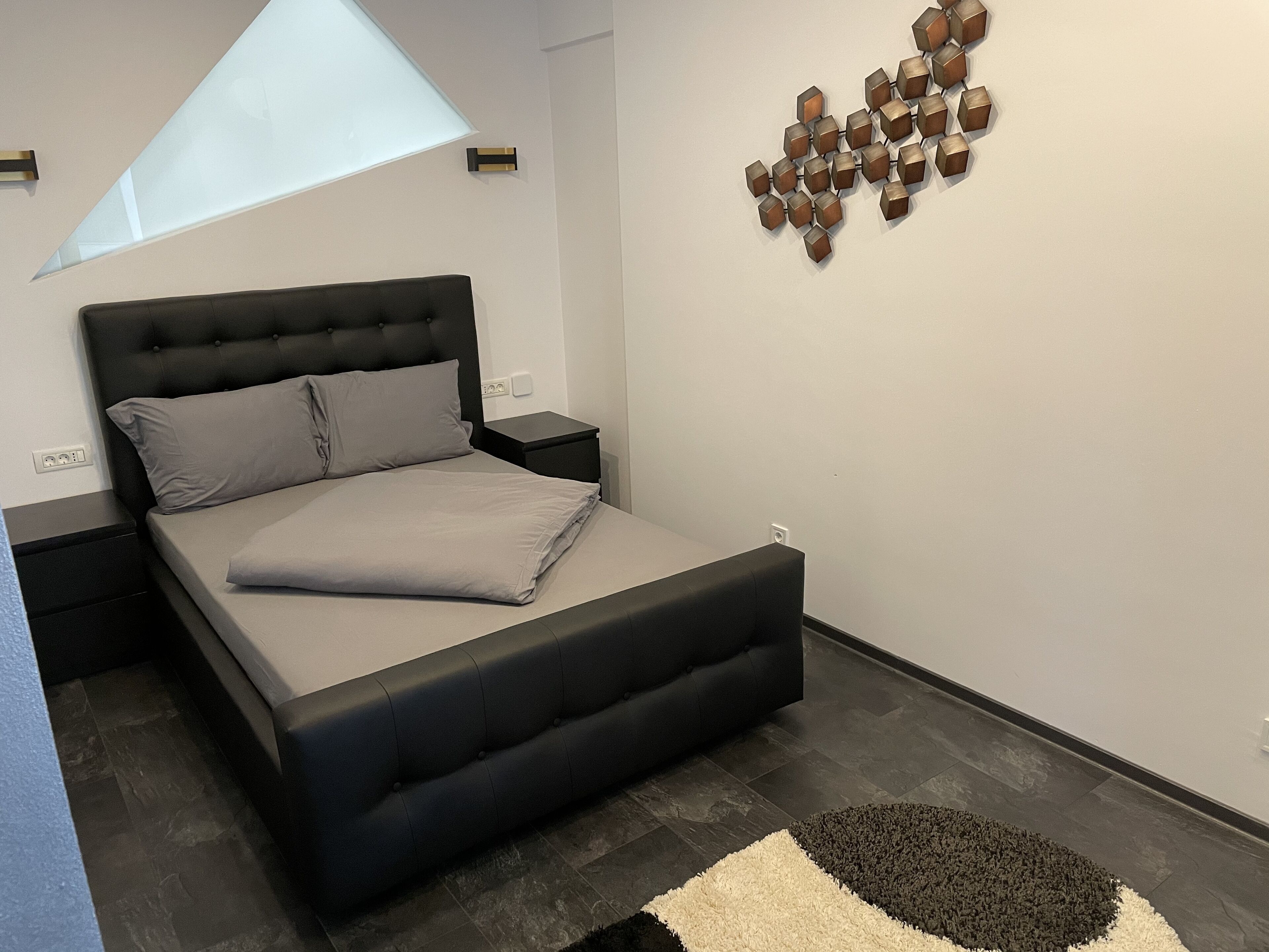 Un dormitorio moderno con una cama grande, una mesita de noche, una lámpara de pared y una obra de arte geométrica.