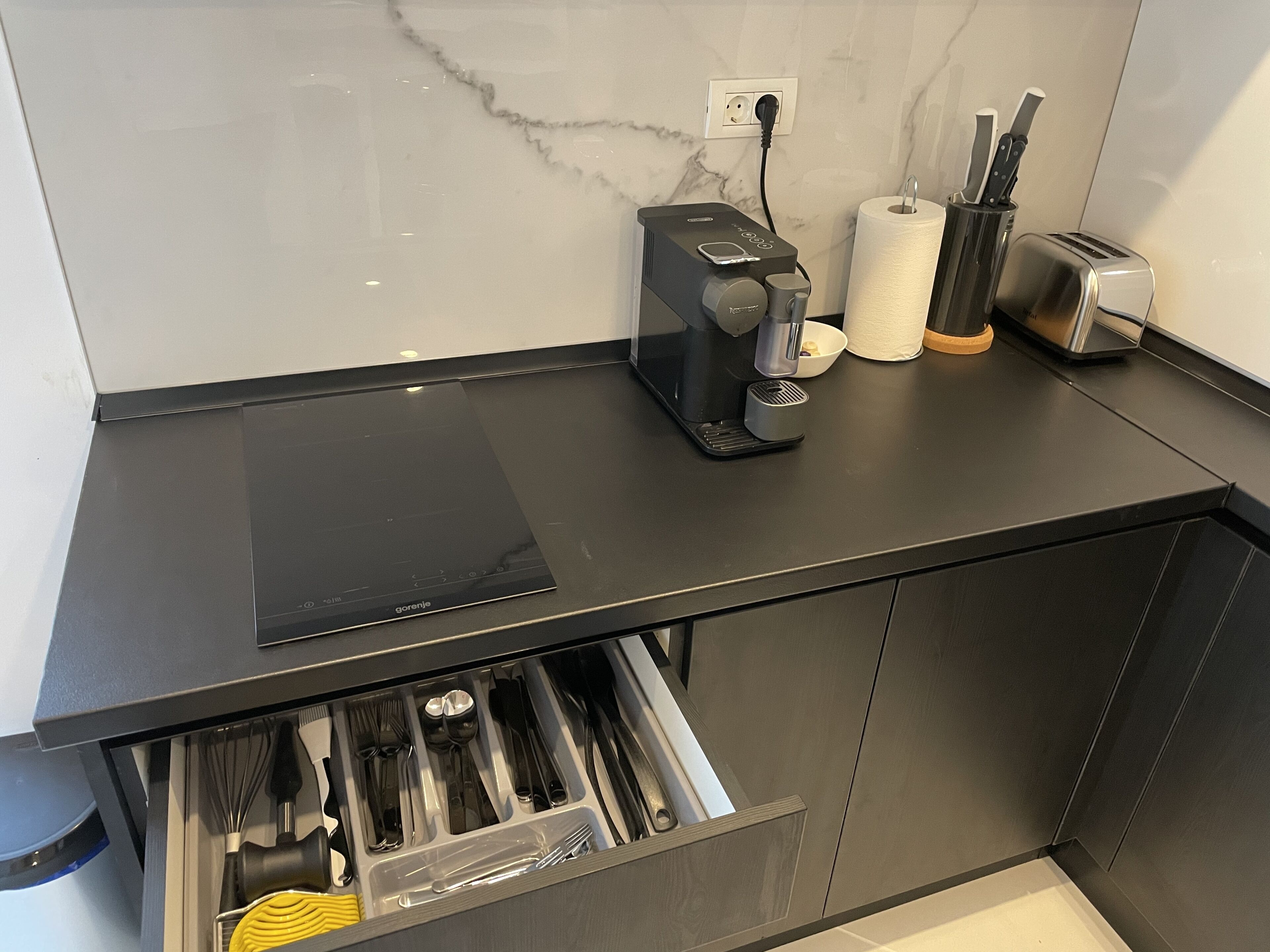 Eine moderne Küche mit schwarzem Arbeitsplatz, einer Kaffeemaschine, einem Toaster und einer Schublade mit Besteck.