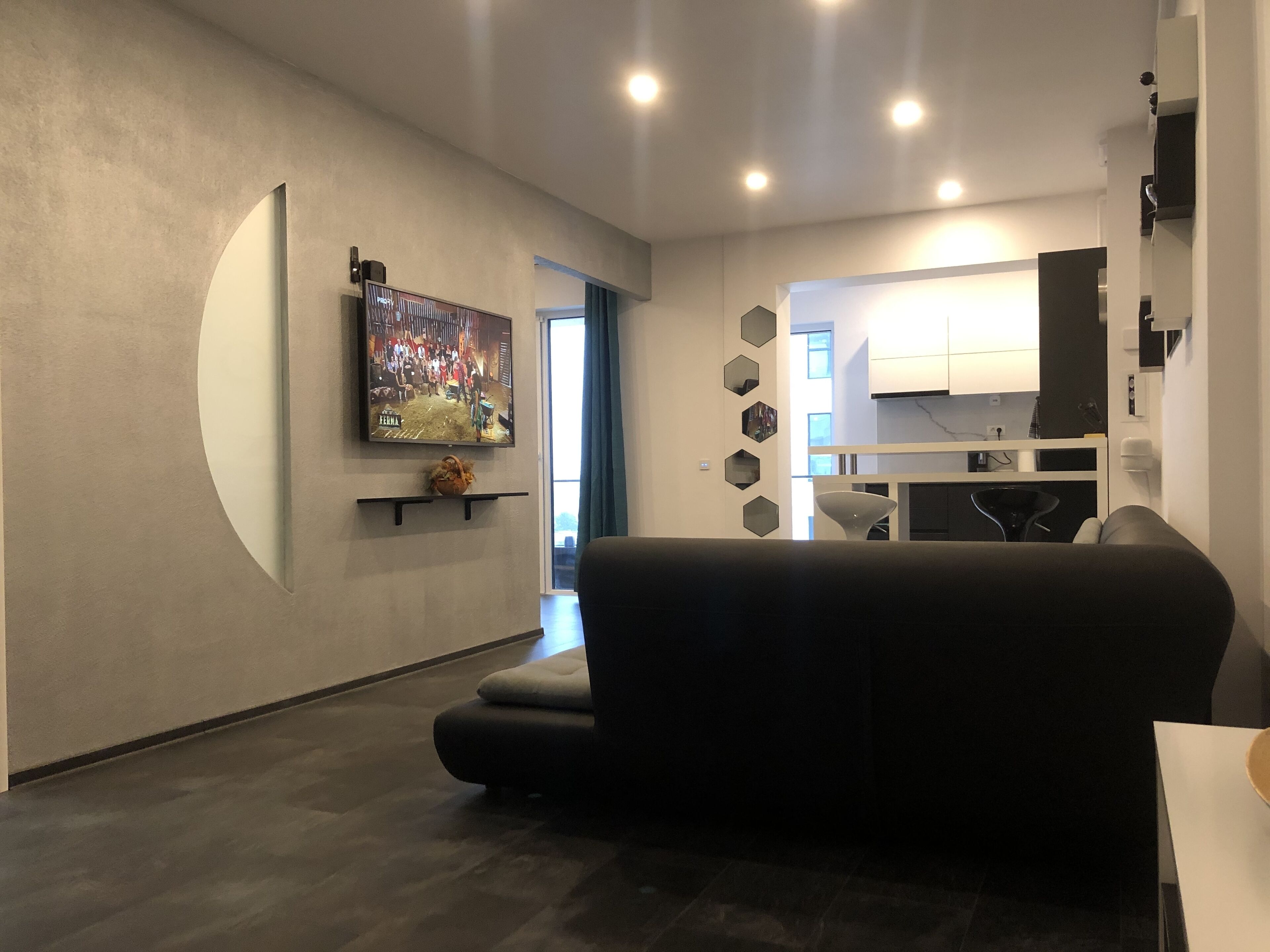 A modern living room with a black sofa, a flat-screen TV mounted on the wall, and a kitchen area visible through an open doorway.