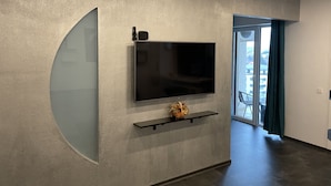 A modern living room with a large flat-screen TV mounted on a textured wall, a small shelf below, and a curved glass partition.