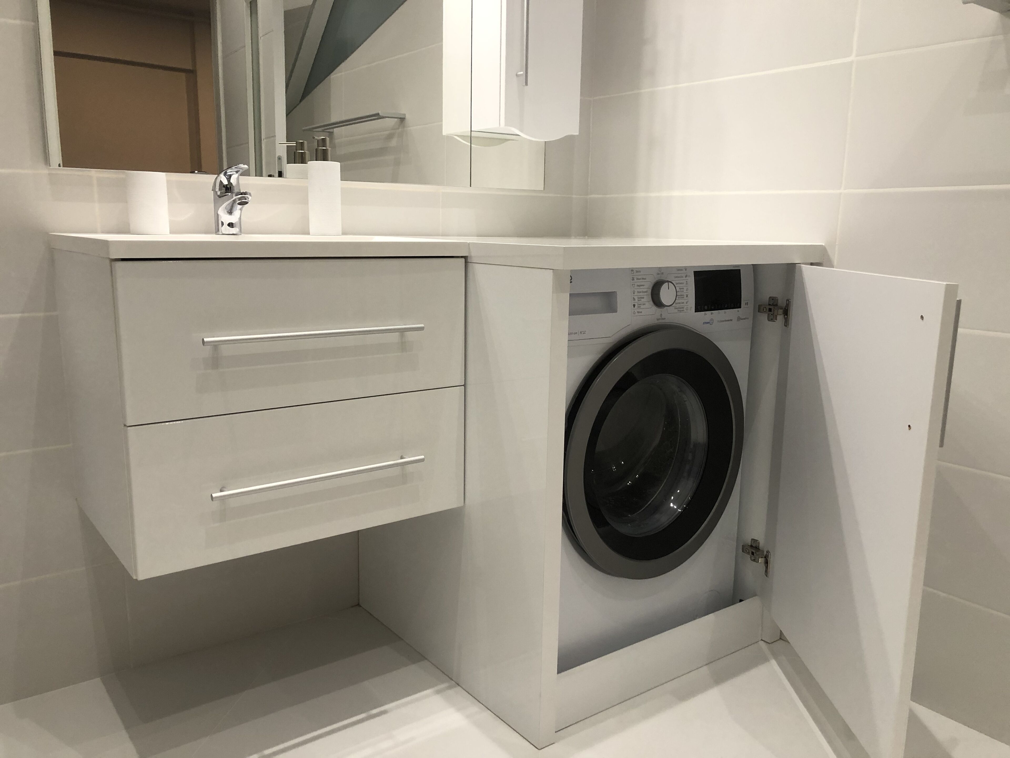 A compact, modern bathroom with a built-in washing machine, a sink with a mirror, and a storage cabinet.