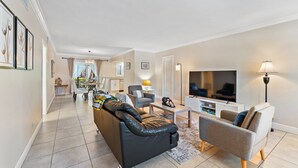 Ein modernes Wohnzimmer mit einer schwarzen Ledersofa, grauen Sesseln, einem Flachbildfernseher und einem Essbereich im Hintergrund.