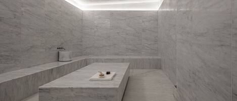 A modern bathroom with a marble floor and walls, a rectangular sink with a towel dispenser, and a small table with a cup and a book.
