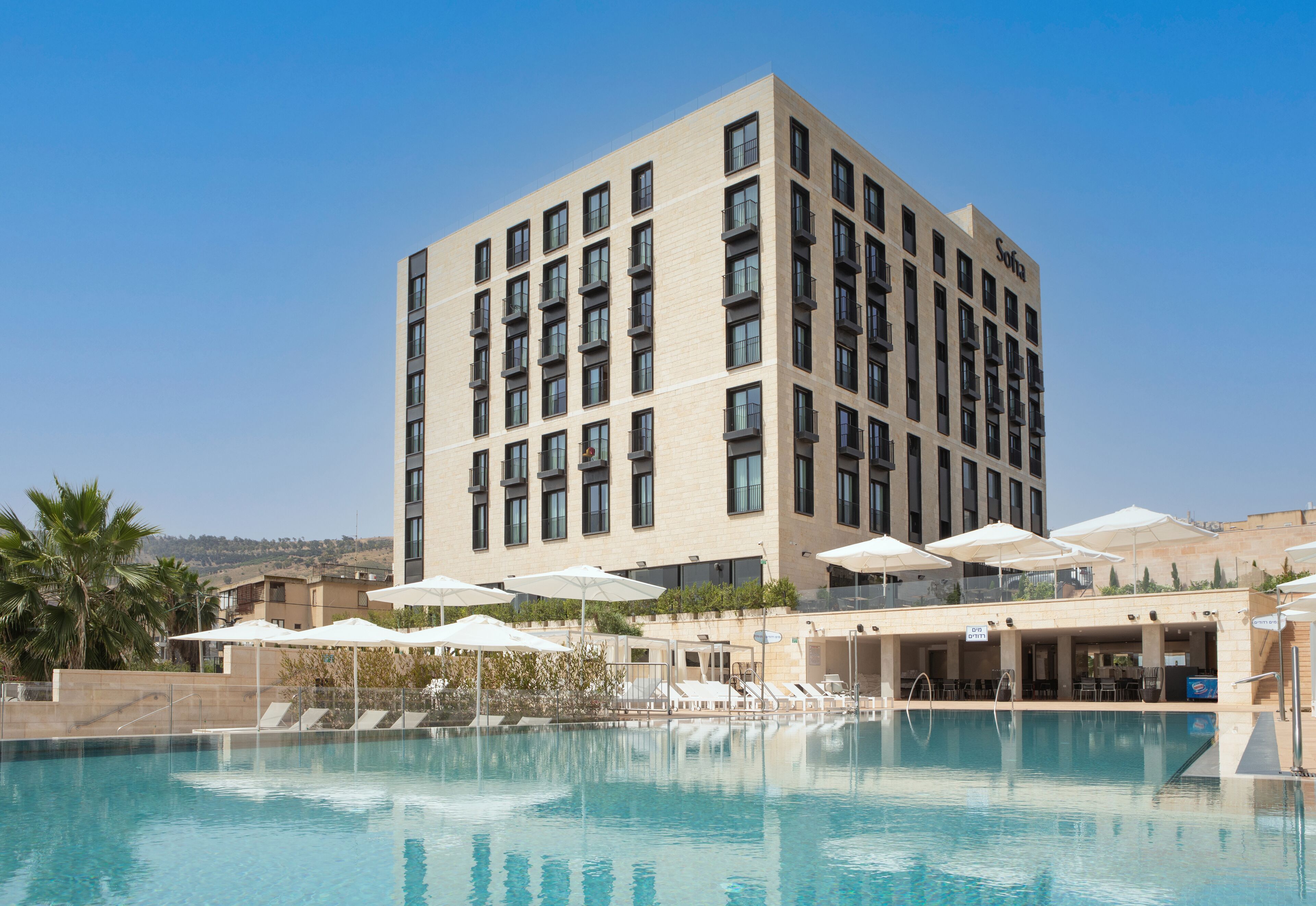 Un hotel moderno con zona de piscina, varios balcones y mobiliario exterior bajo sombrillas.