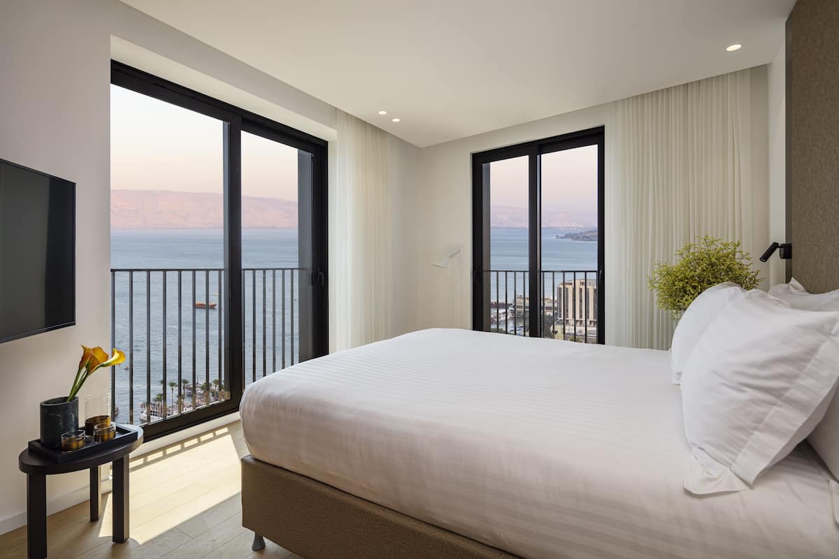 A modern hotel room with a large bed, a TV, and a balcony with a view of the sea.