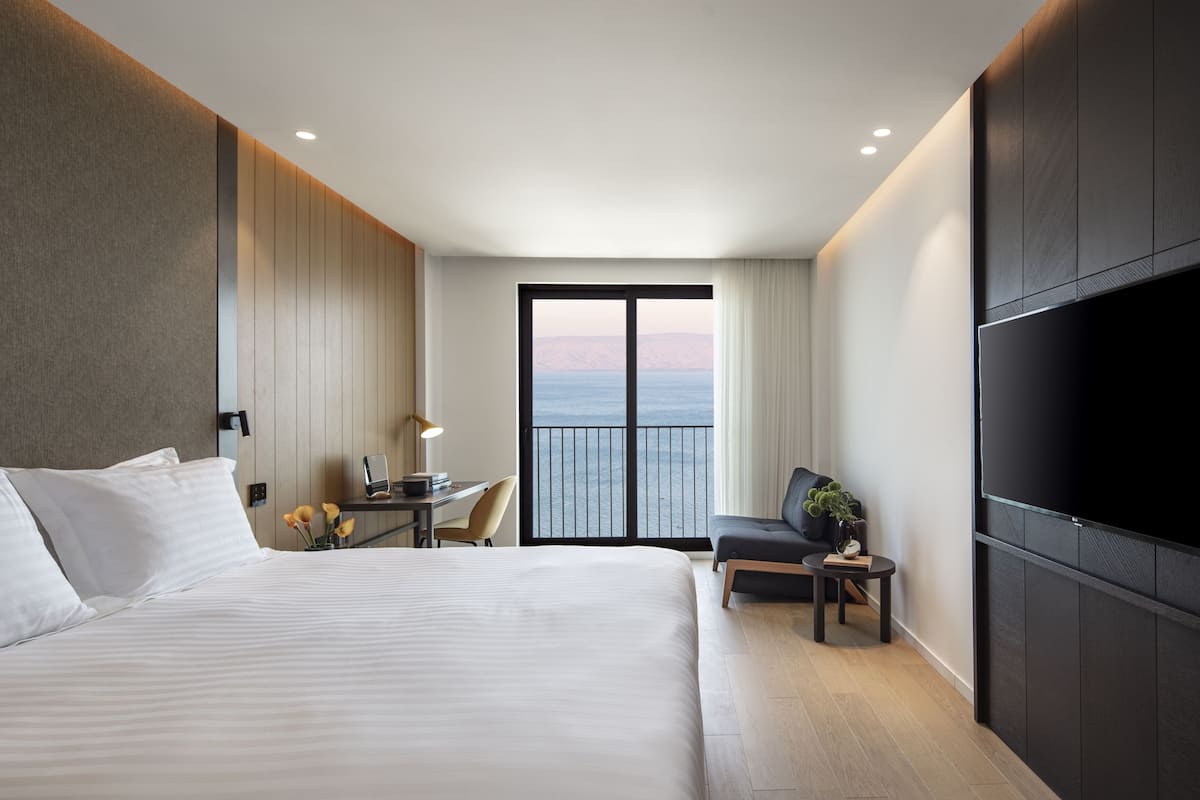 A modern hotel room with a large bed, a desk with a lamp, a chair, a TV, and a balcony with a view of the sea.