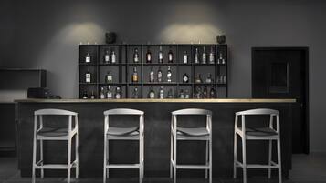 Zona de bar con una pared oscura, estantes llenos de botellas de licor y tres taburetes blancos.