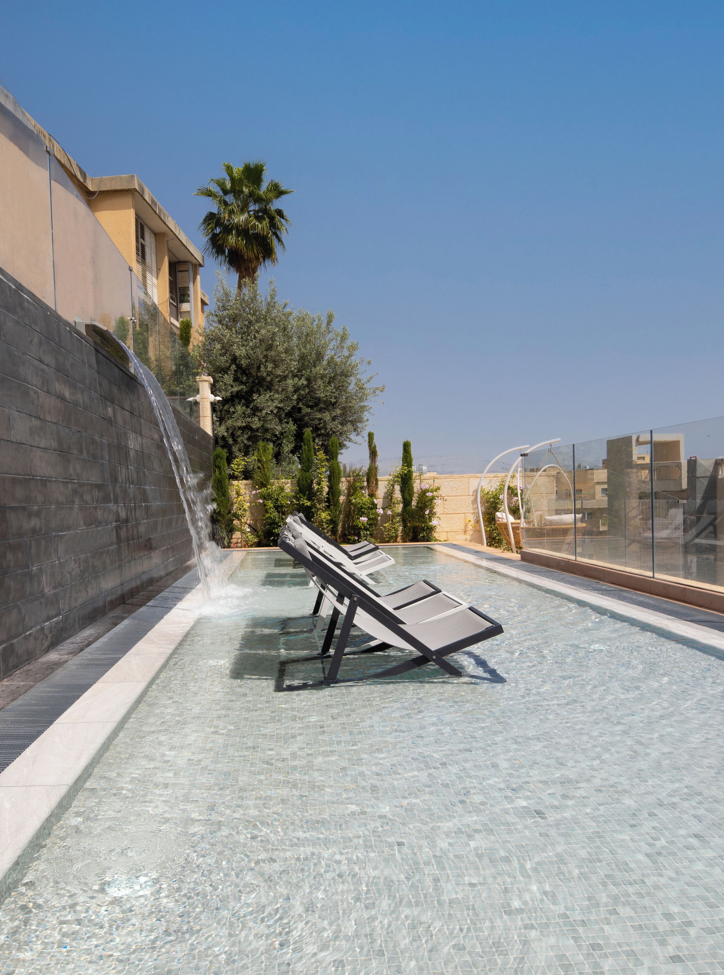 A swimming pool with lounge chairs, a waterfall feature, and a view of buildings and trees.