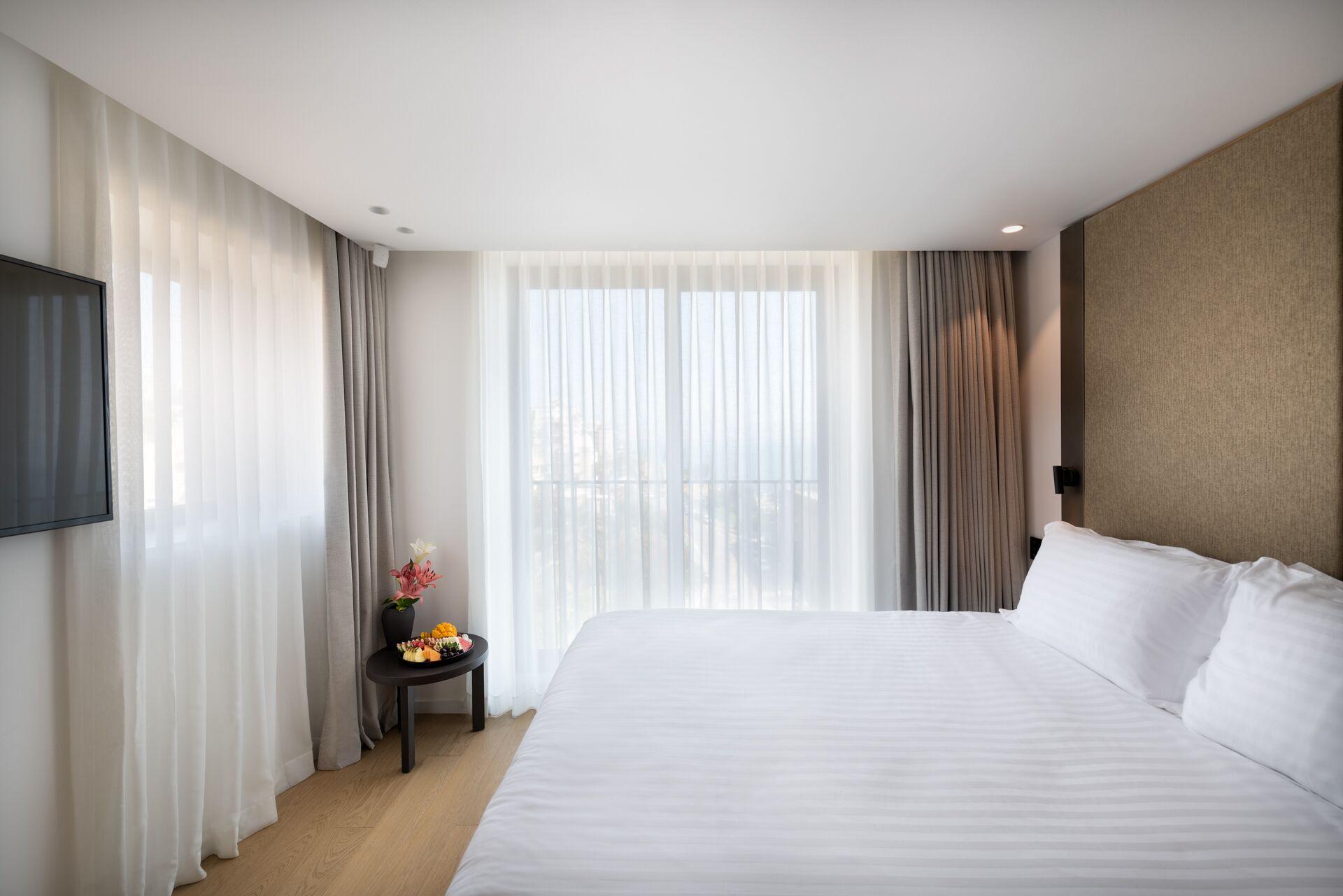 A hotel room with a large bed, a flat-screen TV on the wall, a small table with a vase of flowers, and a window with sheer curtains.