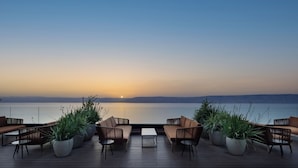 Una terraza con mobiliario y plantas en macetas, con vistas a una masa de agua tranquila al atardecer.
