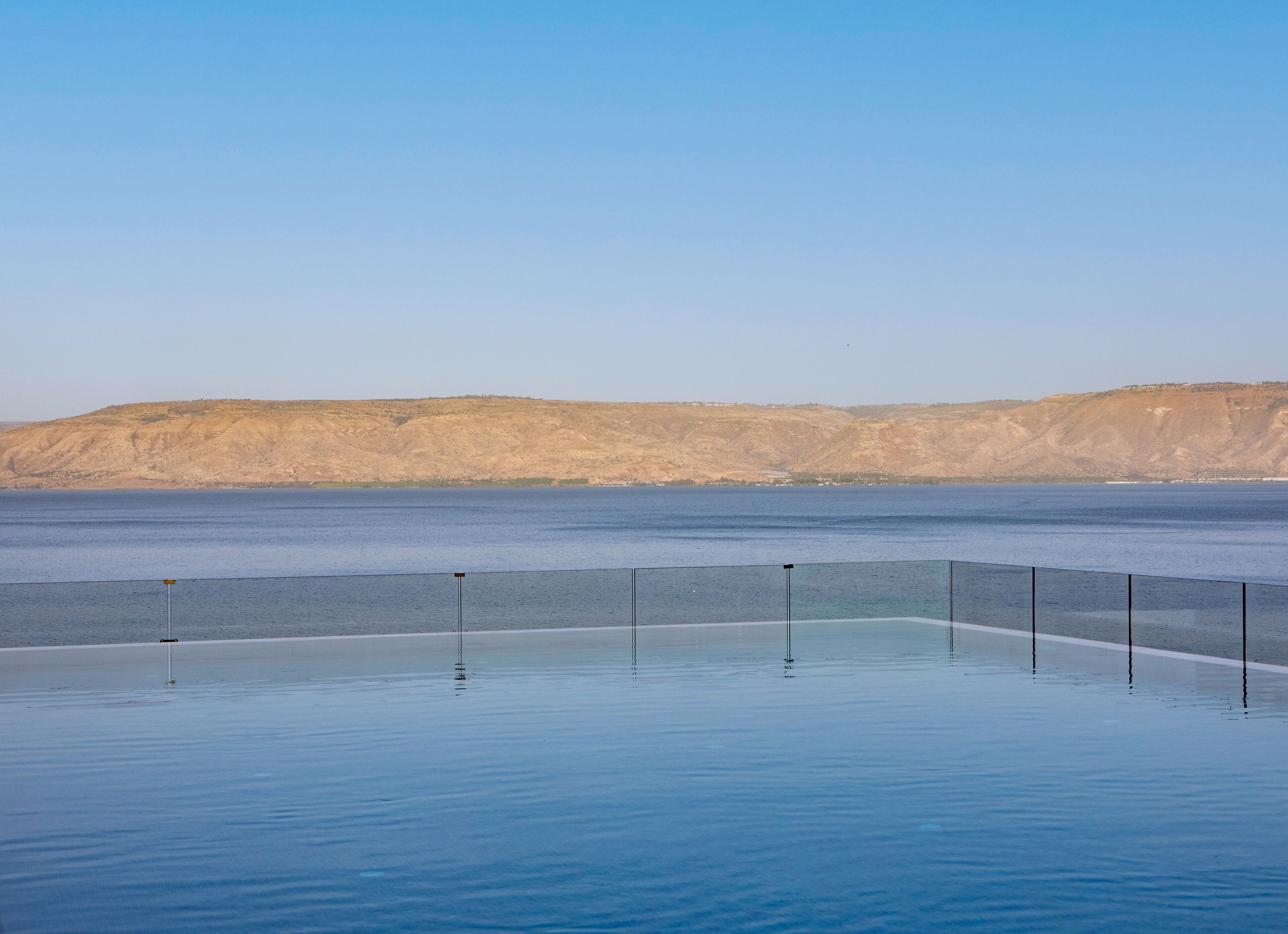 A clear view of a swimming pool with a glass barrier, a distant mountain range, and a calm body of water.