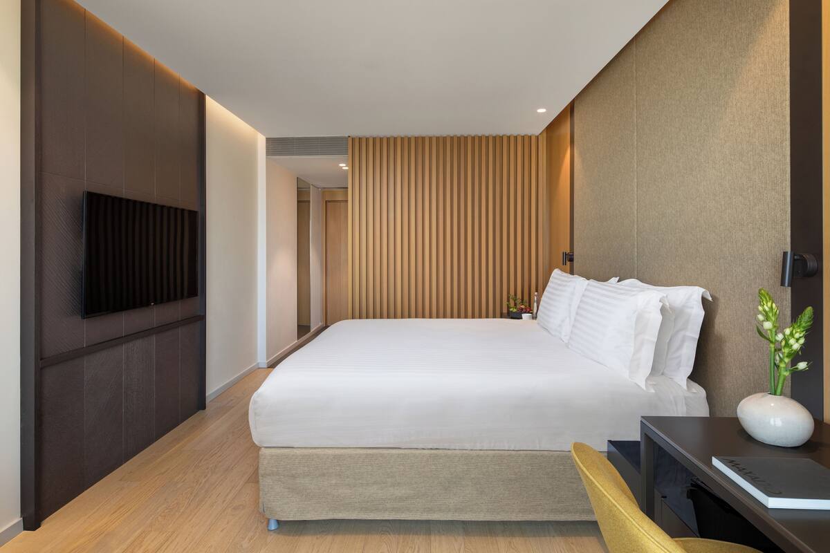A modern hotel room with a large bed, a flat-screen TV mounted on the wall, a bedside table with a vase of flowers, and a wooden panel wall.