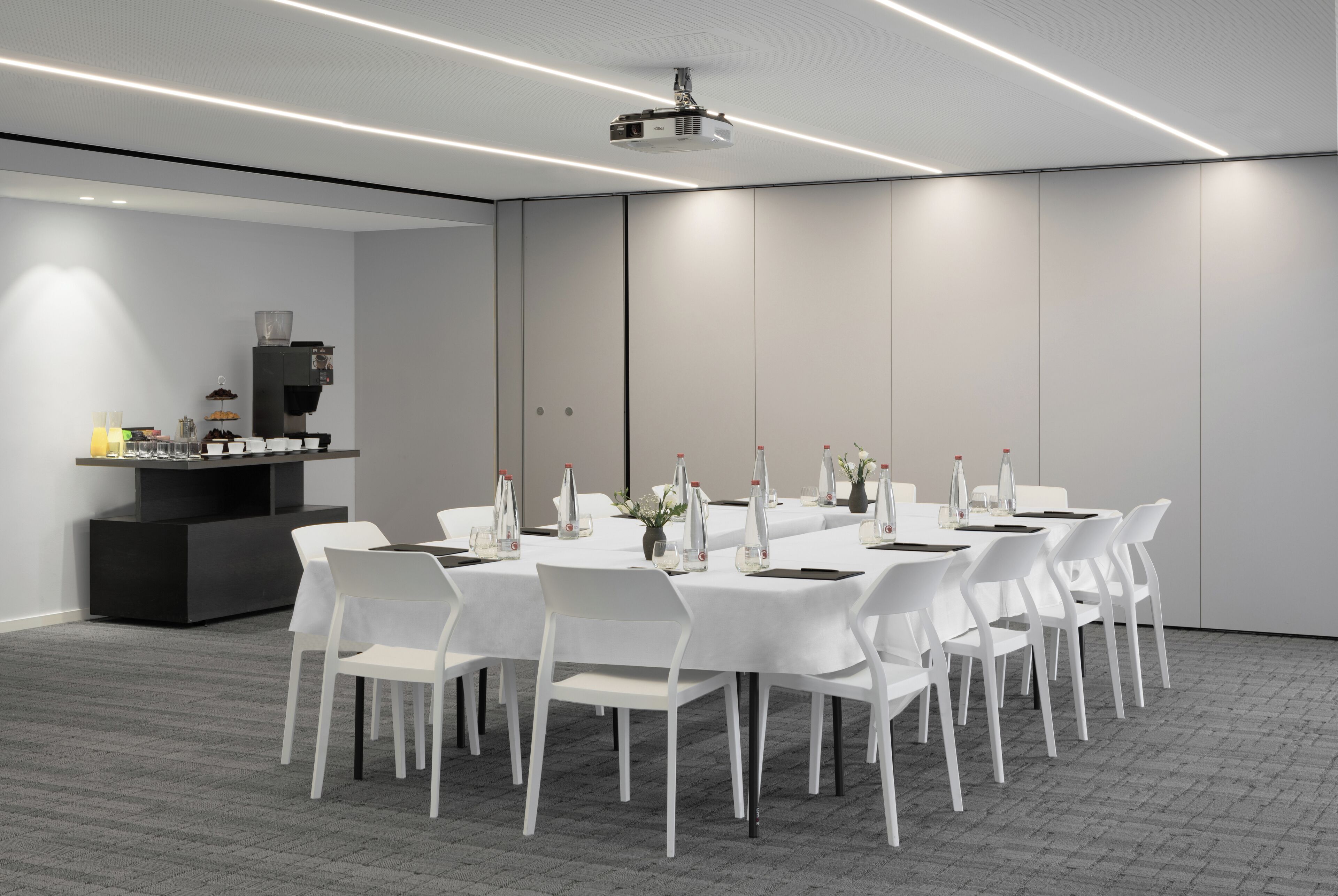 Een vergaderruimte met een lange tafel, witte stoelen en een projector aan het plafond.