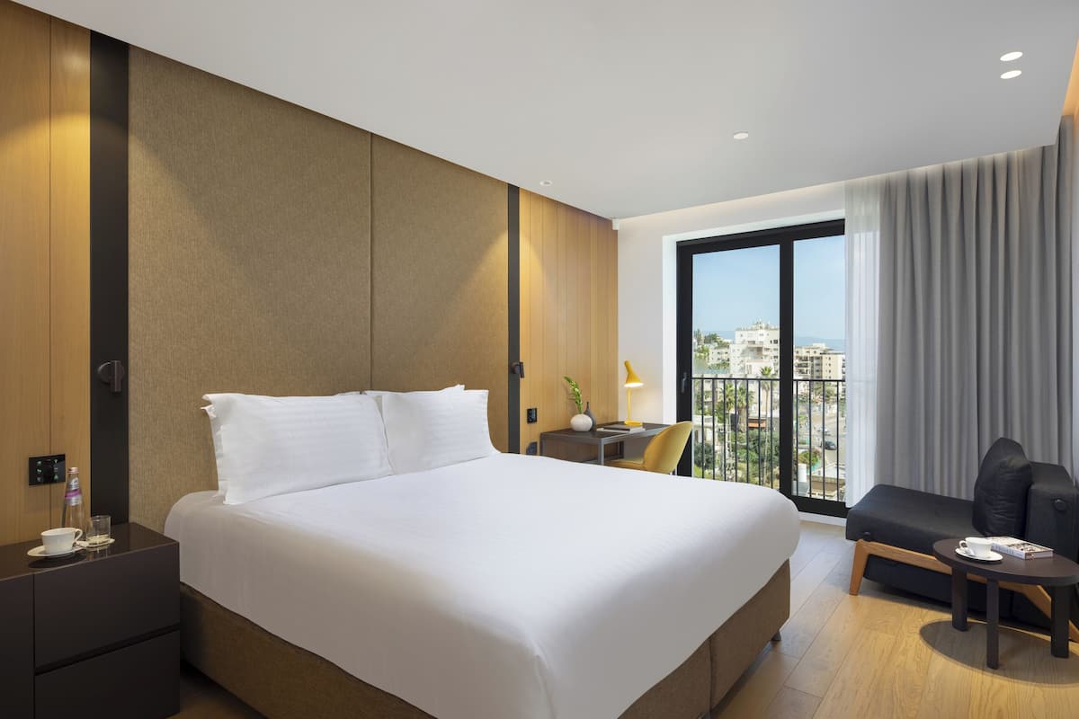 A hotel room with a large bed, a desk with a chair, a small table with a cup and book, a TV, and a balcony with a view of buildings and the sea.