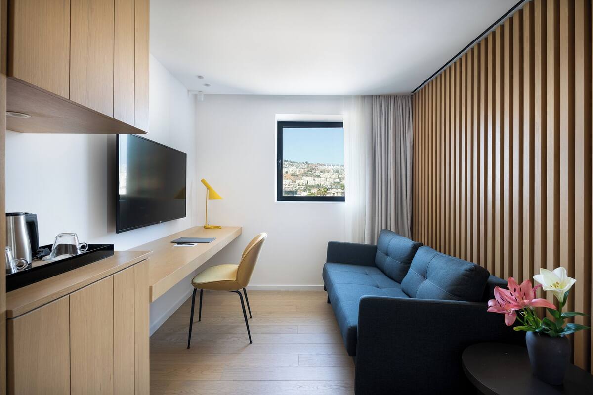 A modern, compact living space with a wooden kitchenette, a flat-screen TV, a desk with a chair, a sofa, and a large window offering a city view.